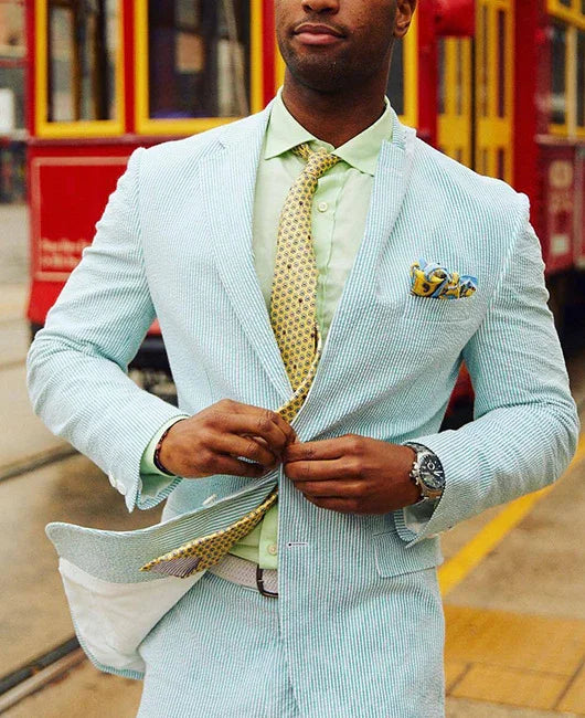 Man in pastel blue seersucker suit with green shirt and patterned yellow tie buttoning jacket near vintage red tram