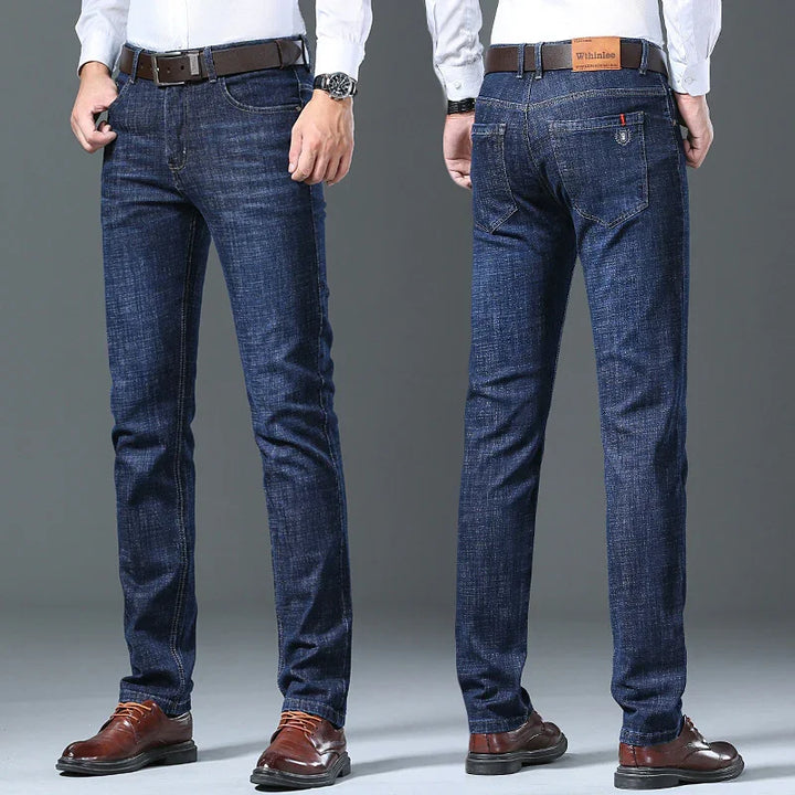 Men's slim fit dark blue jeans styled with brown leather belt and brown dress shoes on gray background