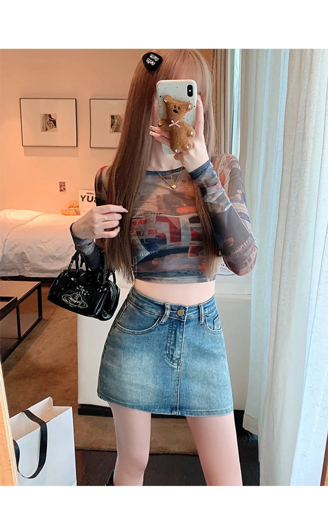 Woman wearing printed mesh crop top and denim mini skirt taking mirror selfie in modern bedroom