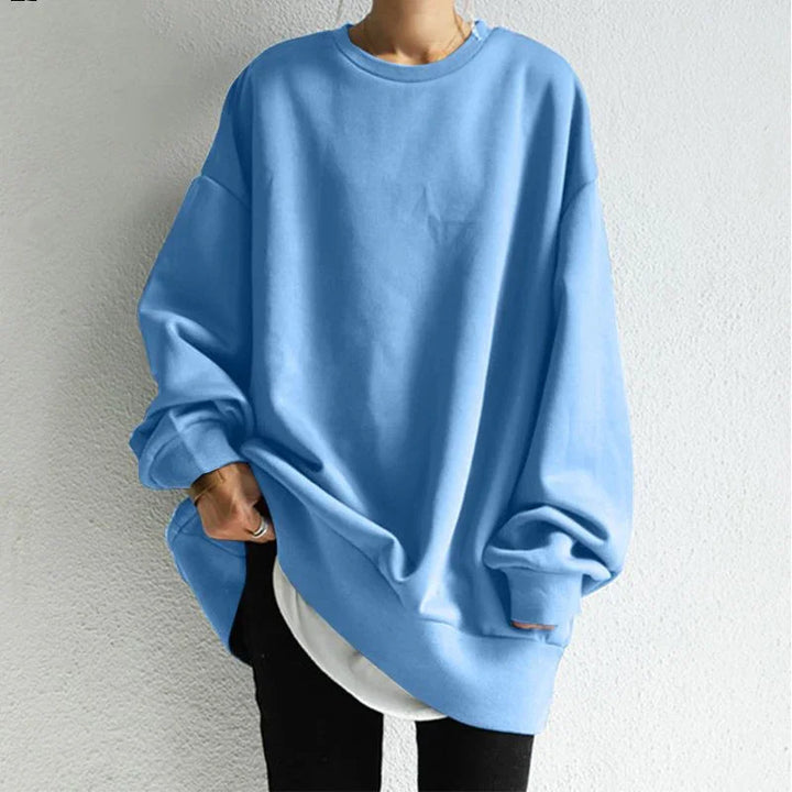 Person wearing oversized light blue sweatshirt with long sleeves and white shirt underneath against white wall