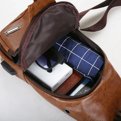 Open brown leather sling bag with charger, folded blue umbrella, phone, and brown wallet inside