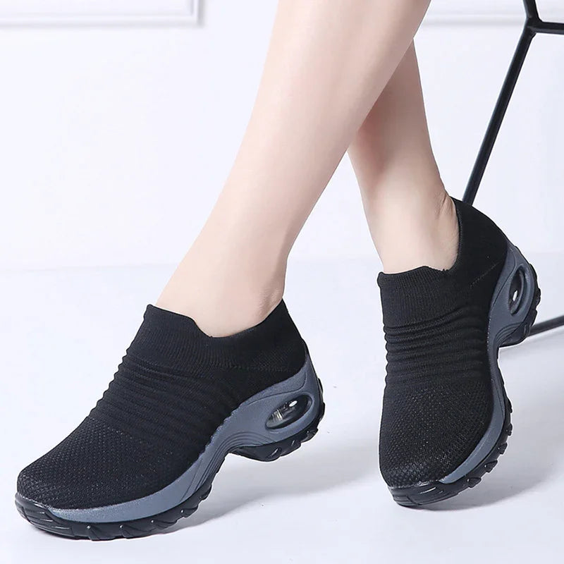 Black slip-on knit sneakers with gray cushioned sole worn on crossed feet