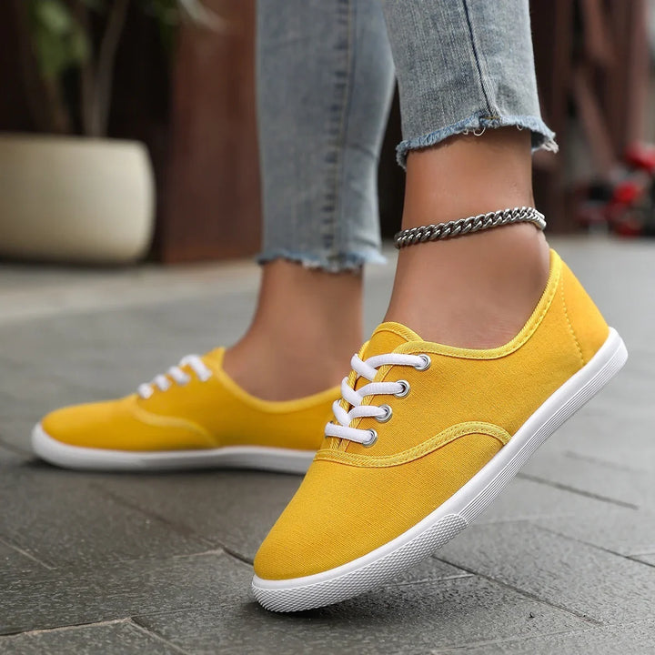 Close-up of feet wearing yellow canvas sneakers with white laces and a silver anklet on grey pavement