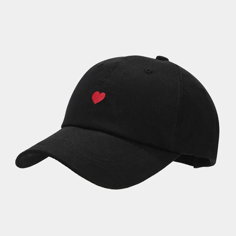 Black baseball cap with small red embroidered heart on front, adjustable strap, plain background