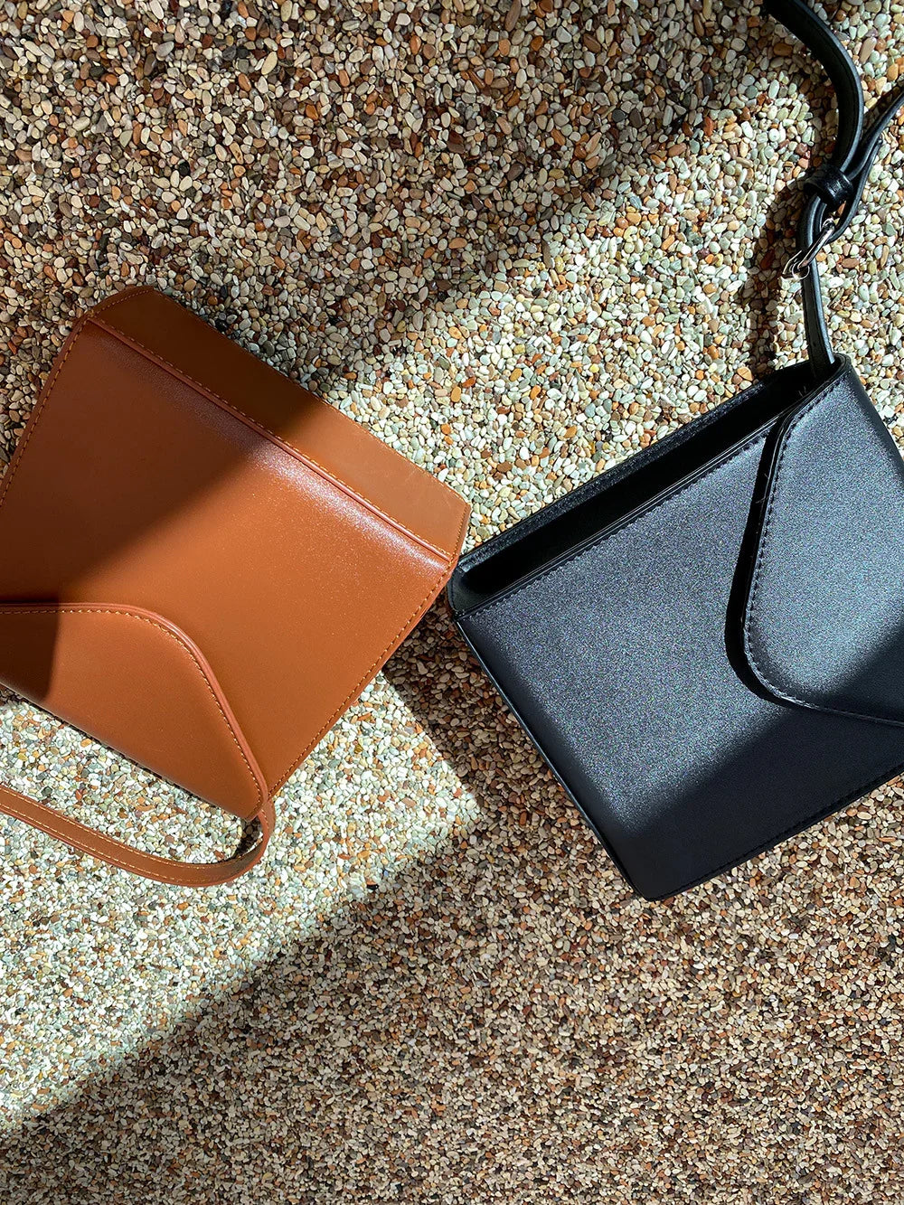 Brown and black leather crossbody bags on a textured pebble surface, lit by natural sunlight