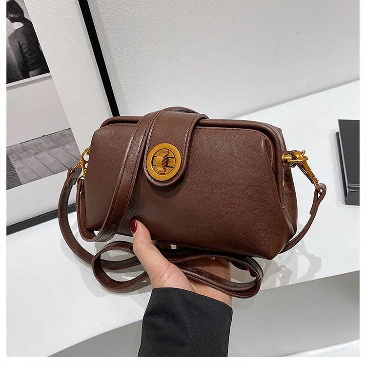 Hand holding a small brown leather crossbody bag with gold clasp and adjustable strap on white surface