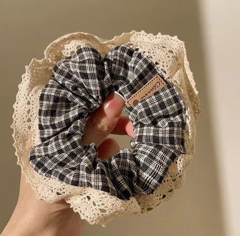 Hand holding a black and white checkered fabric scrunchie with beige lace trim against a plain background