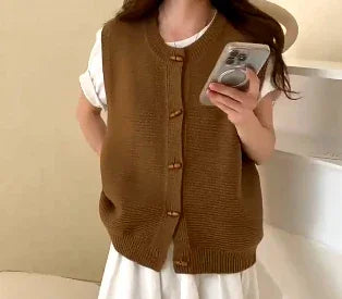 Woman wearing brown knitted buttoned vest over white outfit holding smartphone indoors