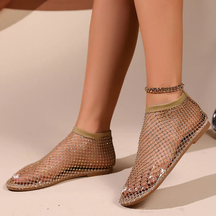 Close-up of feet wearing beige mesh flat shoes adorned with small rhinestones and a gold chain anklet