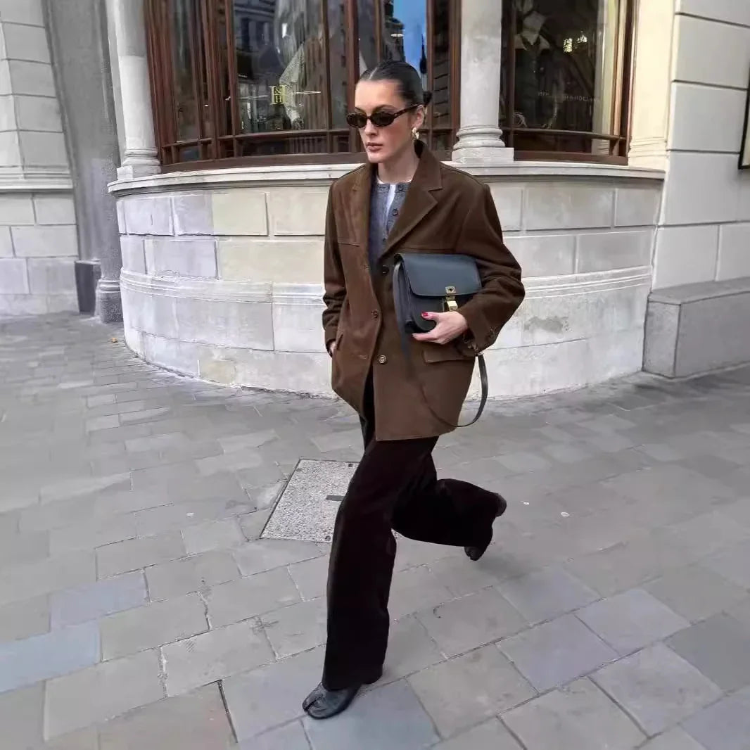 woman wearing brown blazer and black pants walking on city sidewalk holding black handbag