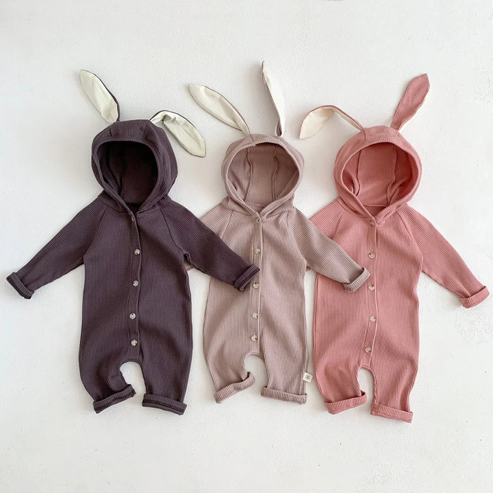 Three ribbed baby onesies with bunny ears hoods in brown, beige, and pink laid on white background