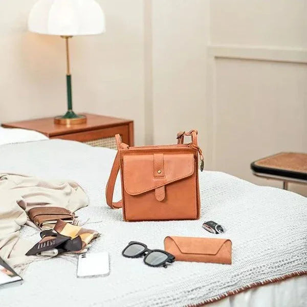 Tan leather crossbody bag on white bed with sunglasses, wallet, keys, and clothing in cozy bedroom