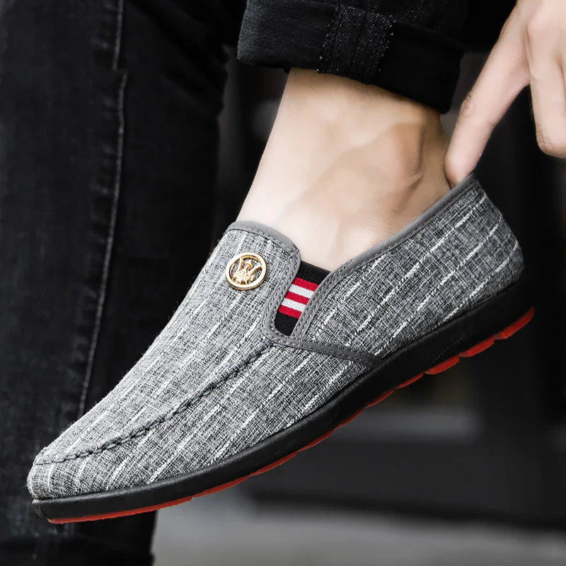 Stylish grey slip-on men's casual shoe with textured fabric, decorative emblem, and black and red sole