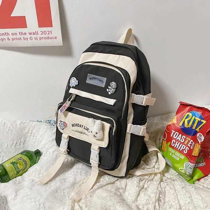 Black and beige Monday Lucy backpack with pins, next to Ritz Toasted Chips and a green bottle on a fabric surface