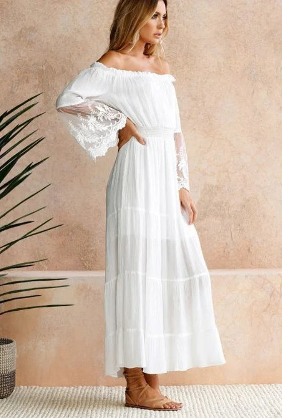 Woman wearing white off-shoulder maxi dress with lace bell sleeves and brown sandals by textured wall