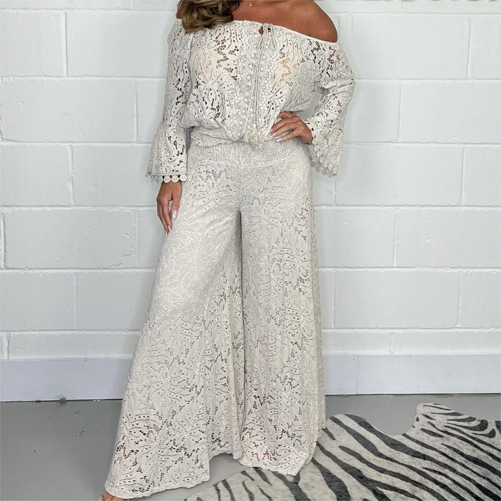 Woman wearing off-shoulder long sleeve white lace jumpsuit with wide legs against white brick wall