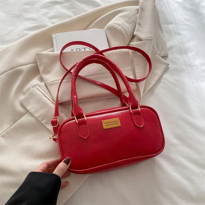 Red leather handbag with gold label and adjustable strap on beige blazer and white bedspread