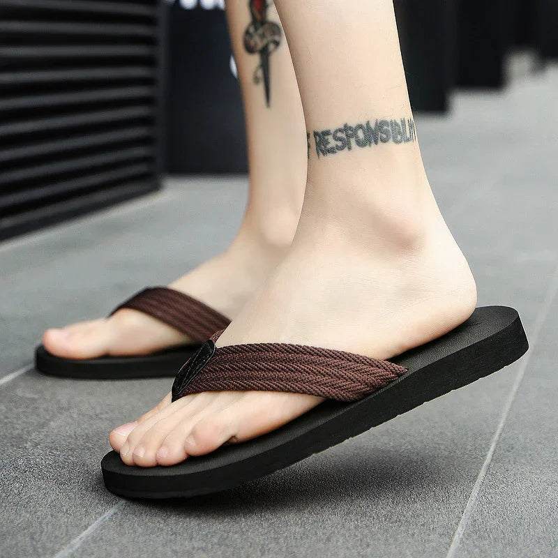 Close-up of feet wearing brown fabric strap flip-flops on gray tiled floor with ankle tattoo