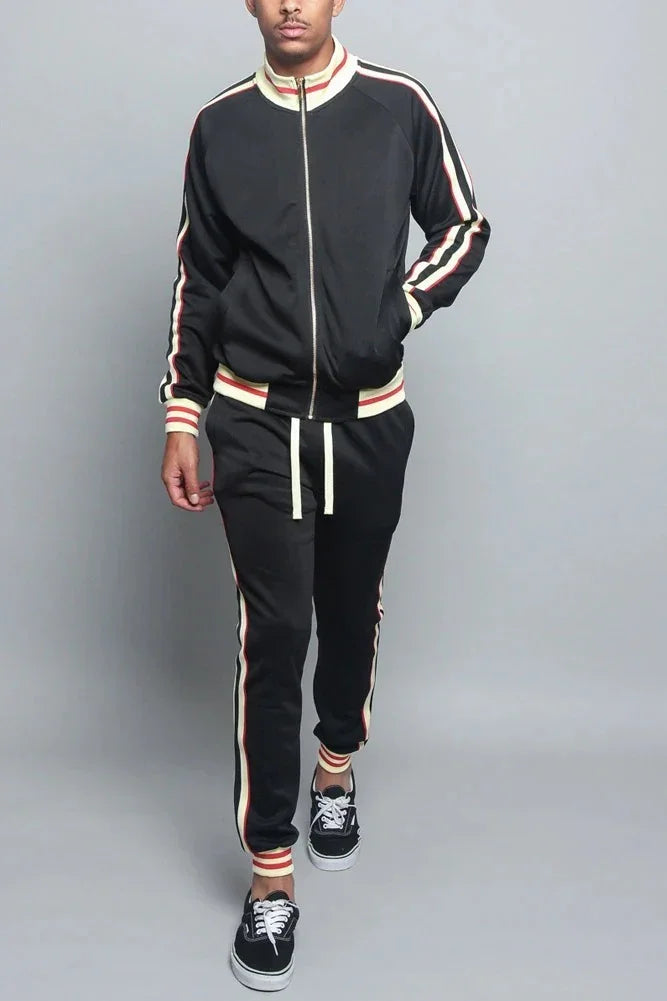 Man modeling black track suit with red and yellow striped trim, casual sneakers, gray background