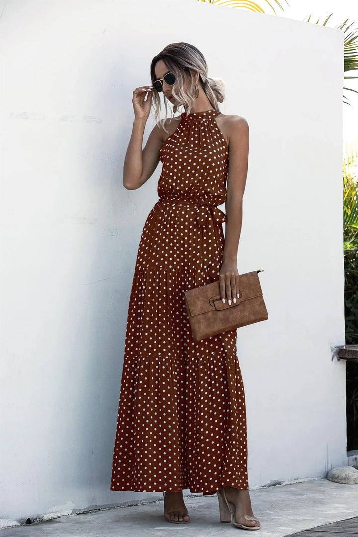 Woman wearing brown polka dot halter maxi dress with belt, holding brown clutch, and sunglasses