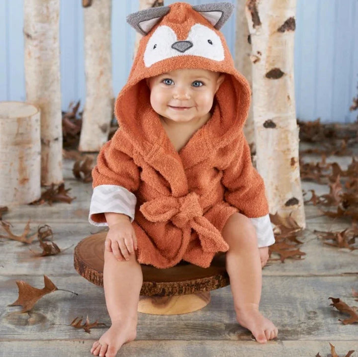 Smiling baby wearing an orange fox hooded towel robe, sitting on wooden stump in autumn setting