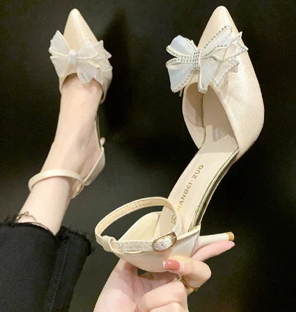 Elegant beige pointed-toe heels with ankle straps and embellished bow detail on black background