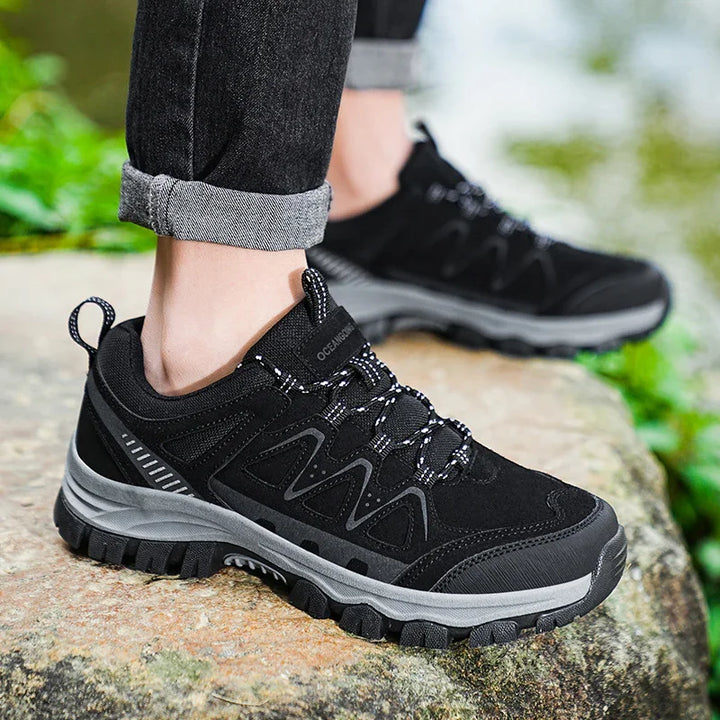 Black hiking shoes with gray soles and patterned laces worn with rolled-up black jeans on a stone surface outdoors