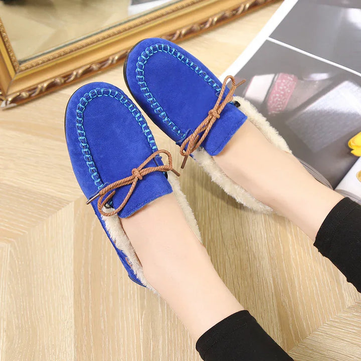 Blue suede moccasin slippers with fur lining and brown bow laces worn indoors on wooden floor