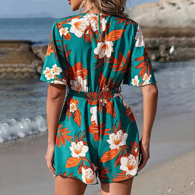 Woman wearing green floral tropical romper with orange and white flowers standing on sandy beach