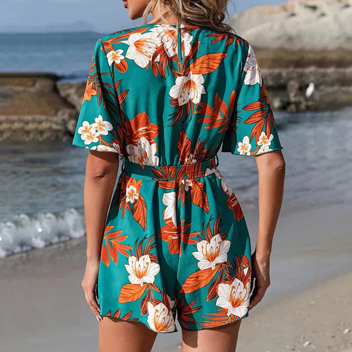 Woman wearing green floral tropical romper with orange and white flowers standing on sandy beach
