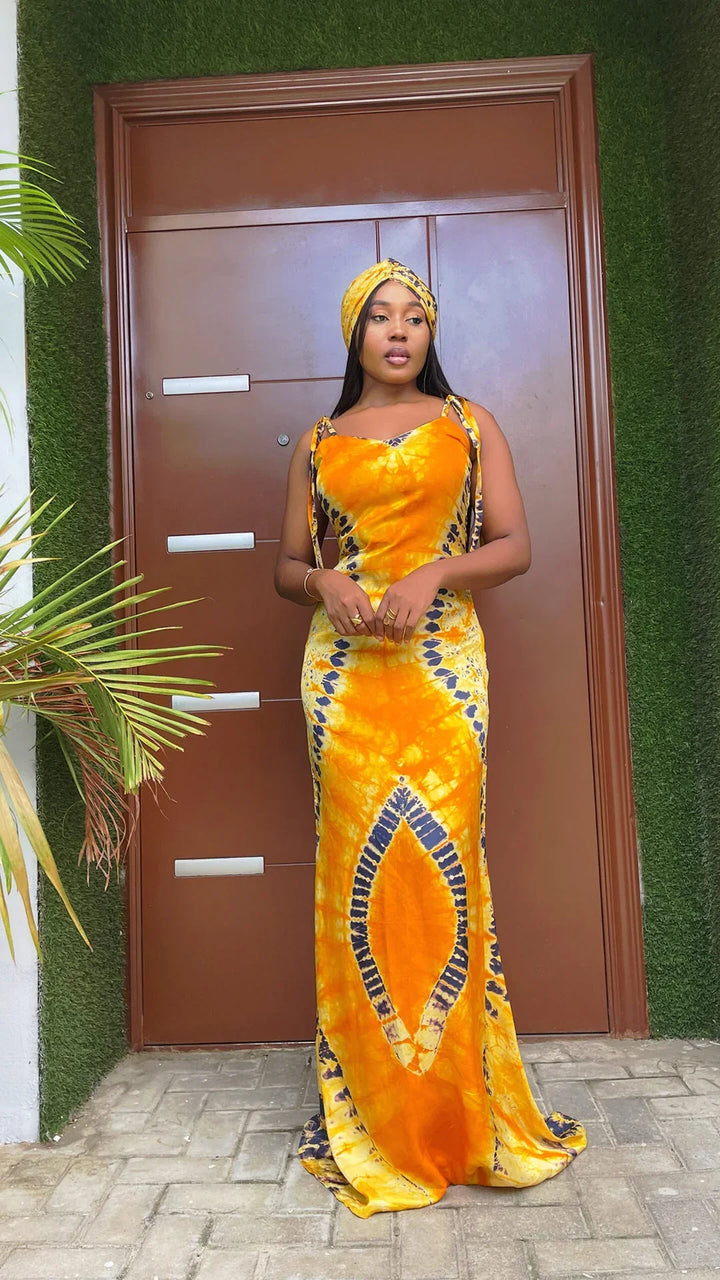 Woman in vibrant yellow and blue patterned maxi dress and matching headwrap standing by brown door with green decor