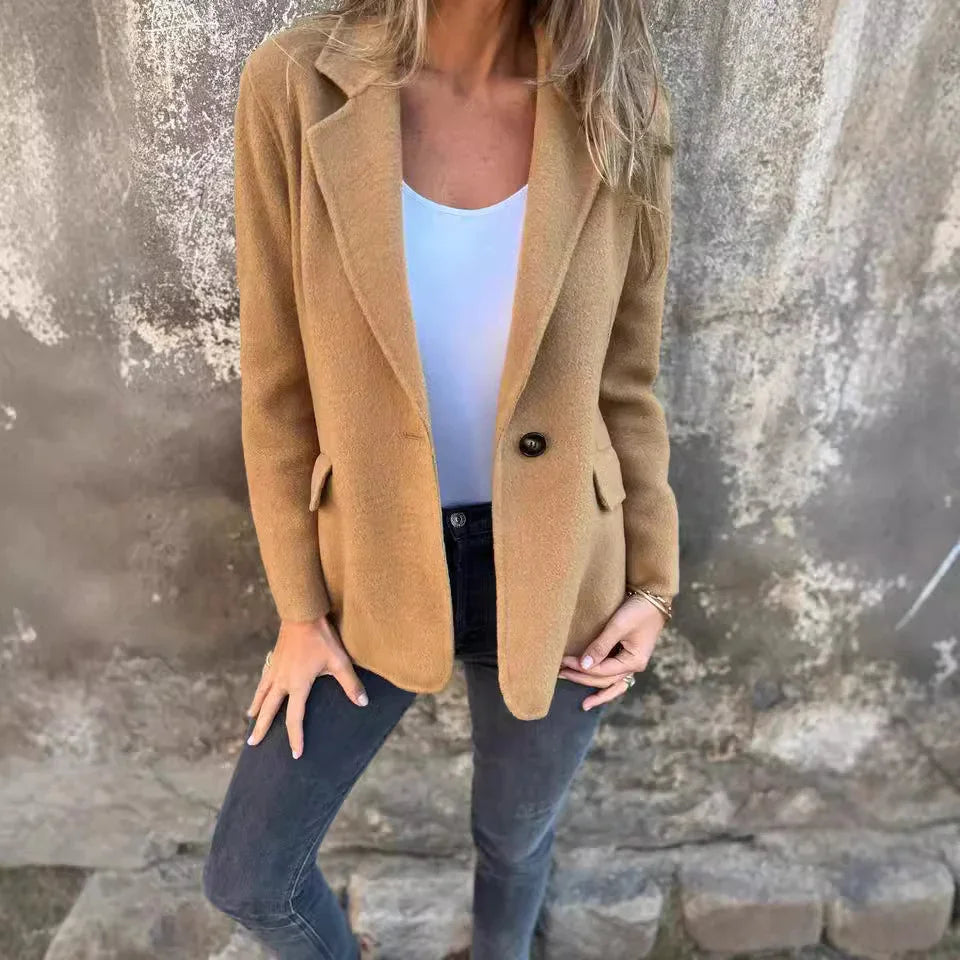 Woman wearing tan blazer with single button, white top, and dark jeans against textured wall