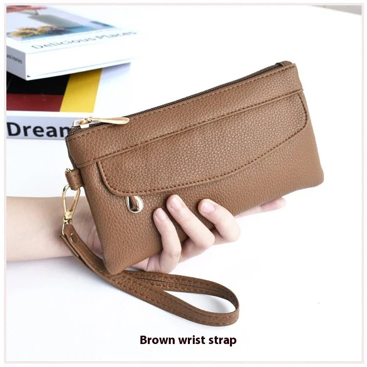 Hand holding a brown textured leather wrist strap clutch purse with gold zipper and metal hooks