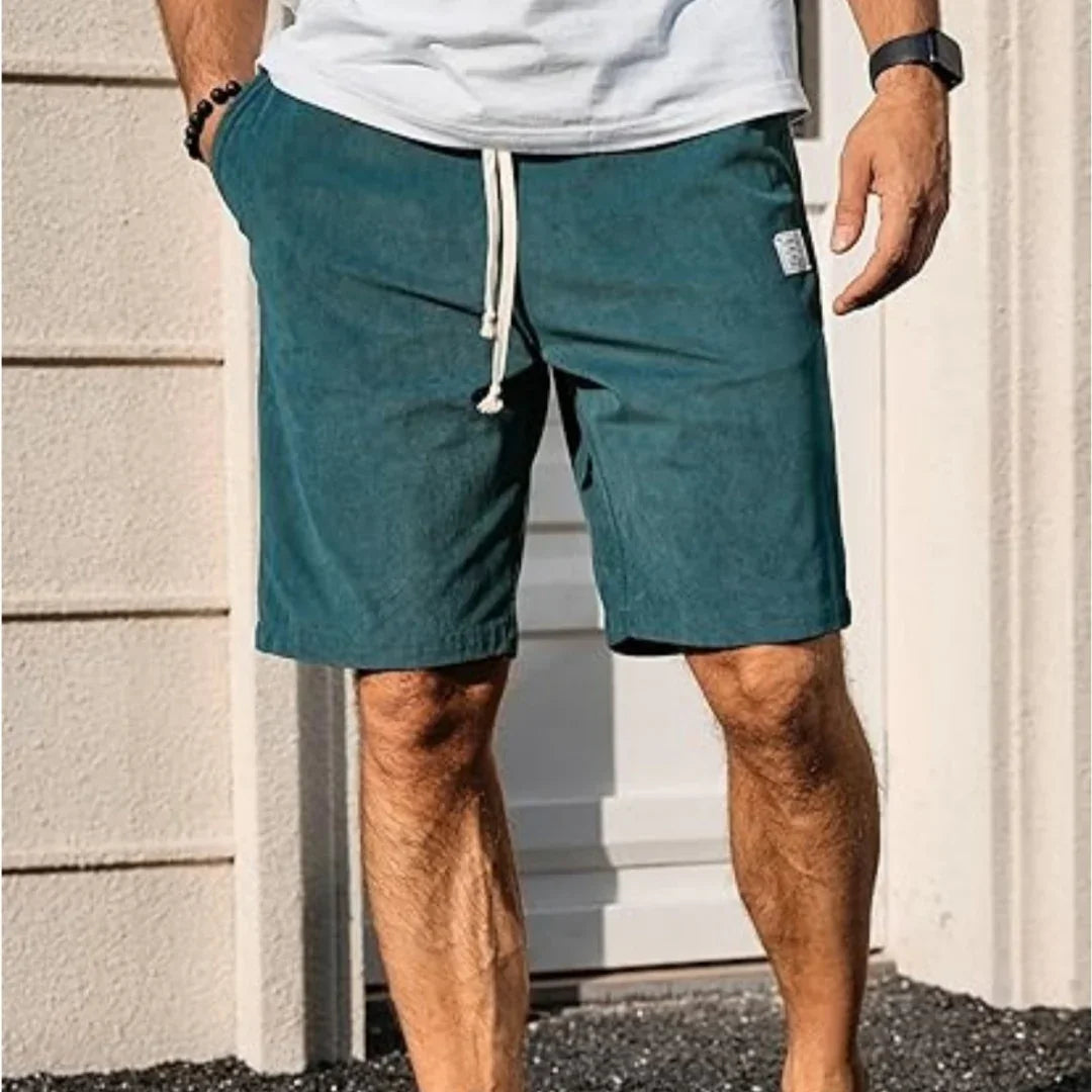 Man wearing teal drawstring shorts and white t-shirt standing outside near a beige wall and door