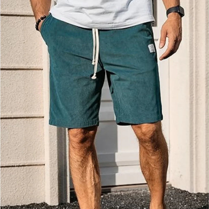 Man wearing teal drawstring shorts and white t-shirt standing outside near a beige wall and door
