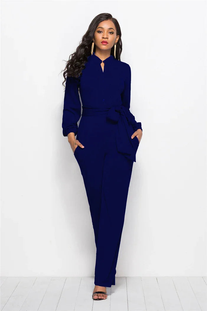 Woman wearing a navy blue long-sleeve jumpsuit with a waist tie and black heels against white background
