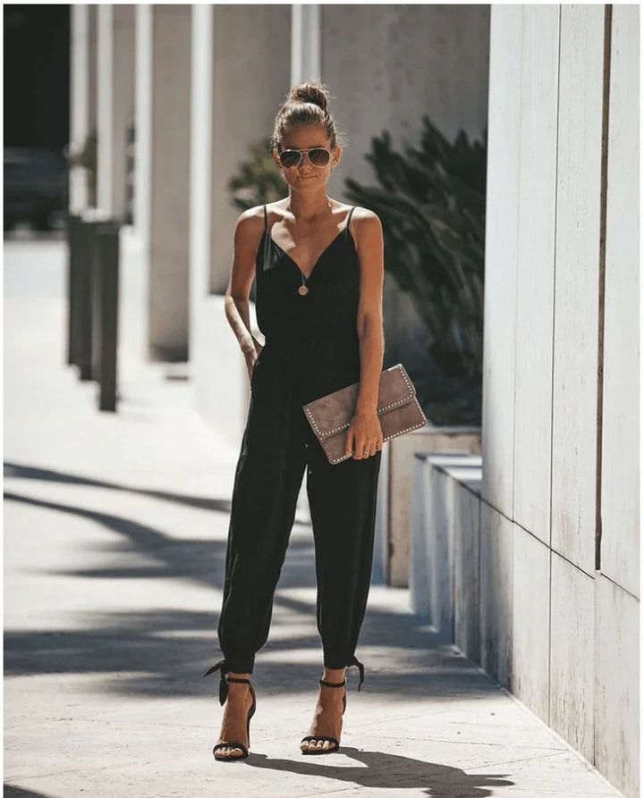 Woman wearing black sleeveless jumpsuit with ankle ties, sunglasses, and holding beige studded clutch outdoors