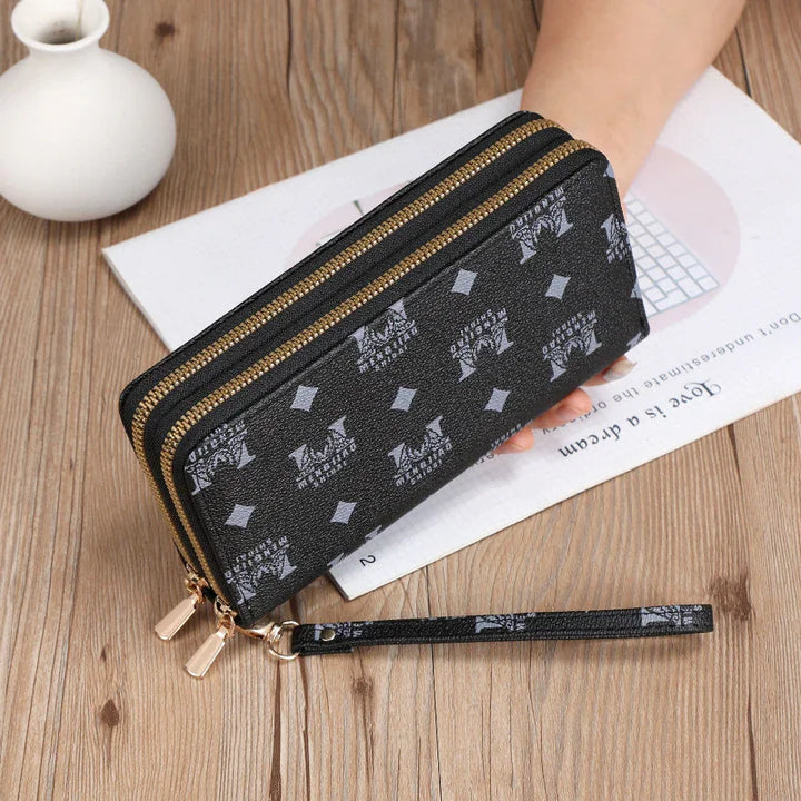 Black leather wristlet wallet with double gold zippers and repeated white logo pattern on wooden table