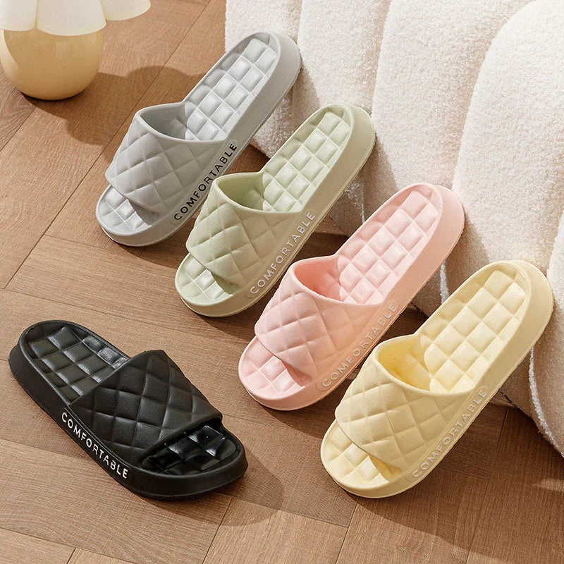 Four pairs of quilted slide sandals in black, gray, green, pink, and yellow on wooden floor with cozy white chair