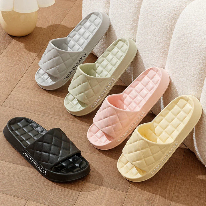 Four pairs of quilted slide sandals in black, gray, green, pink, and yellow on wooden floor with cozy white chair