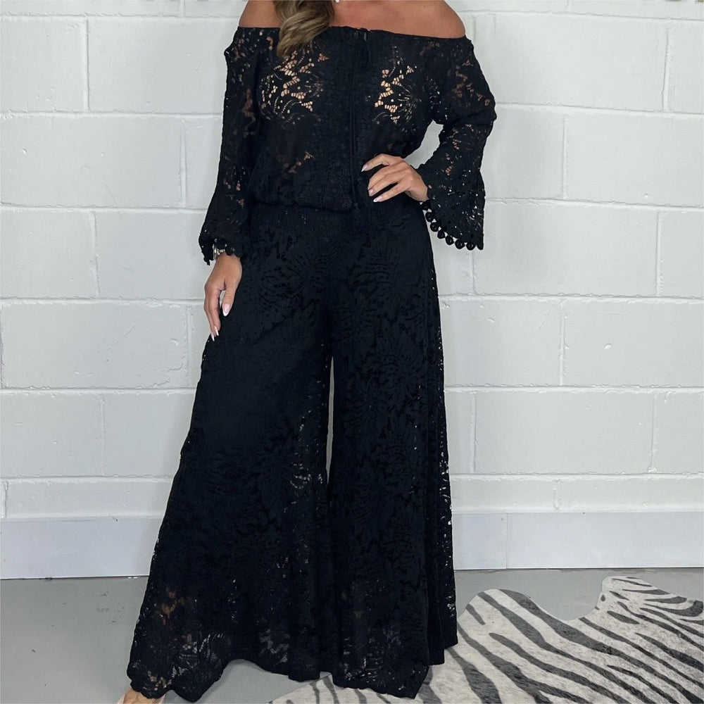 Woman wearing black off-shoulder lace wide-leg jumpsuit against white brick wall