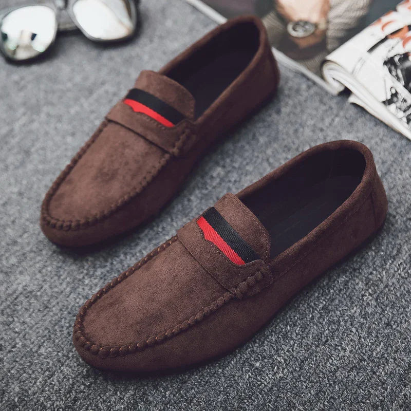 Brown suede loafers with black and red striped detail on gray carpet floor