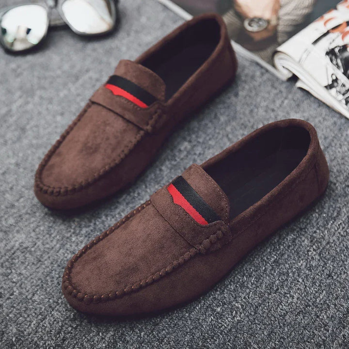 Brown suede loafers with black and red striped detail on gray carpet floor