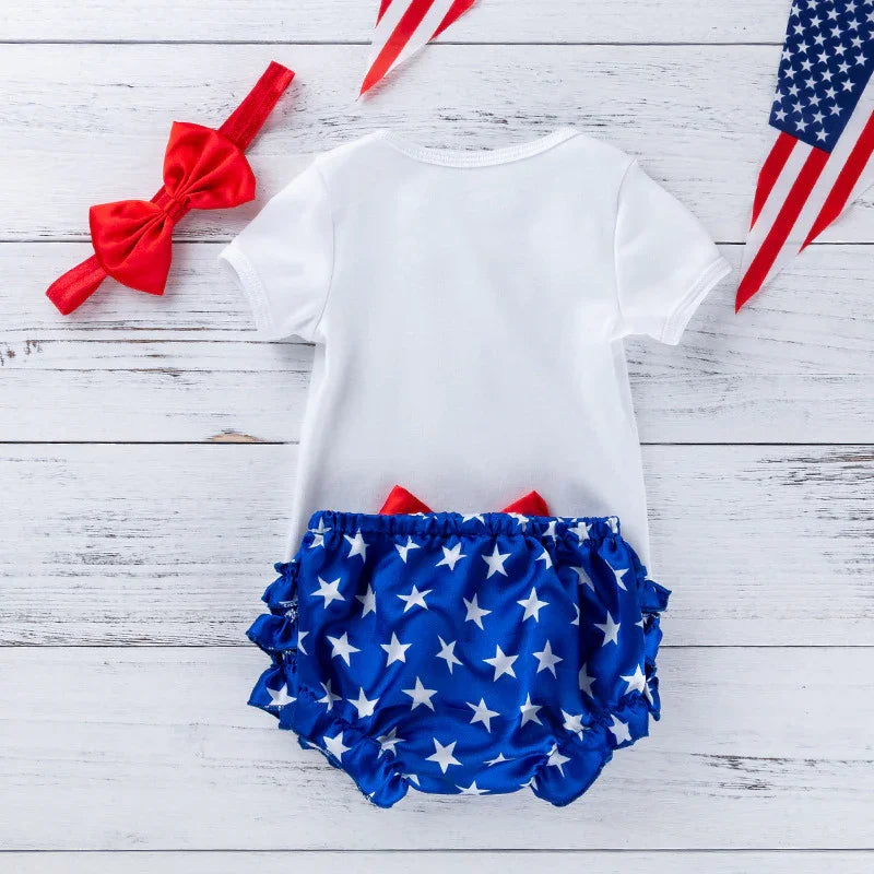 Baby outfit with white short-sleeve shirt, blue star print ruffled bloomers, red bow headband on white wooden background