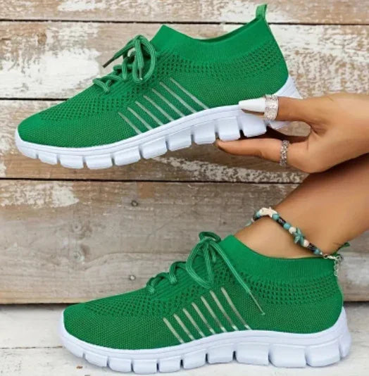 Green knit athletic sneakers with white soles held and worn by hands with rings and a beaded anklet on wooden background
