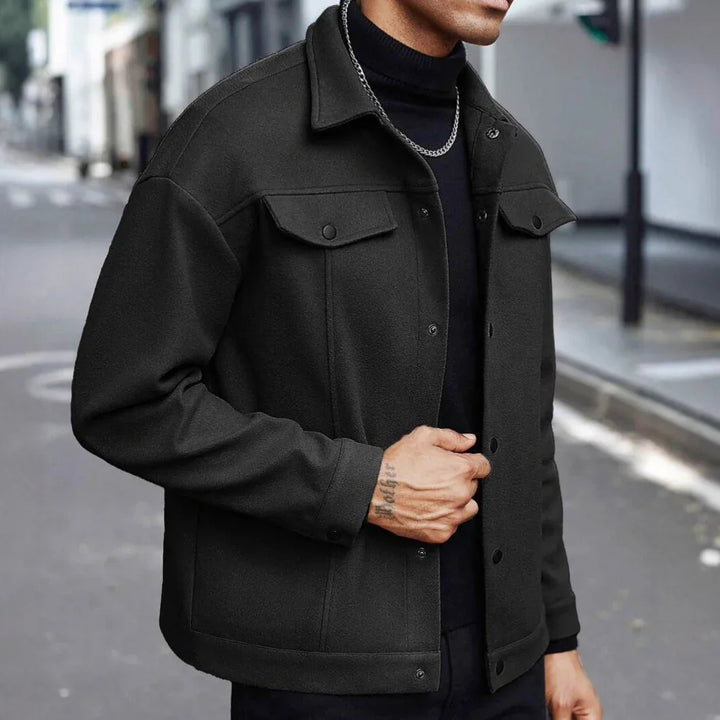 Man wearing a black wool jacket with buttoned chest pockets and a chain necklace on urban street