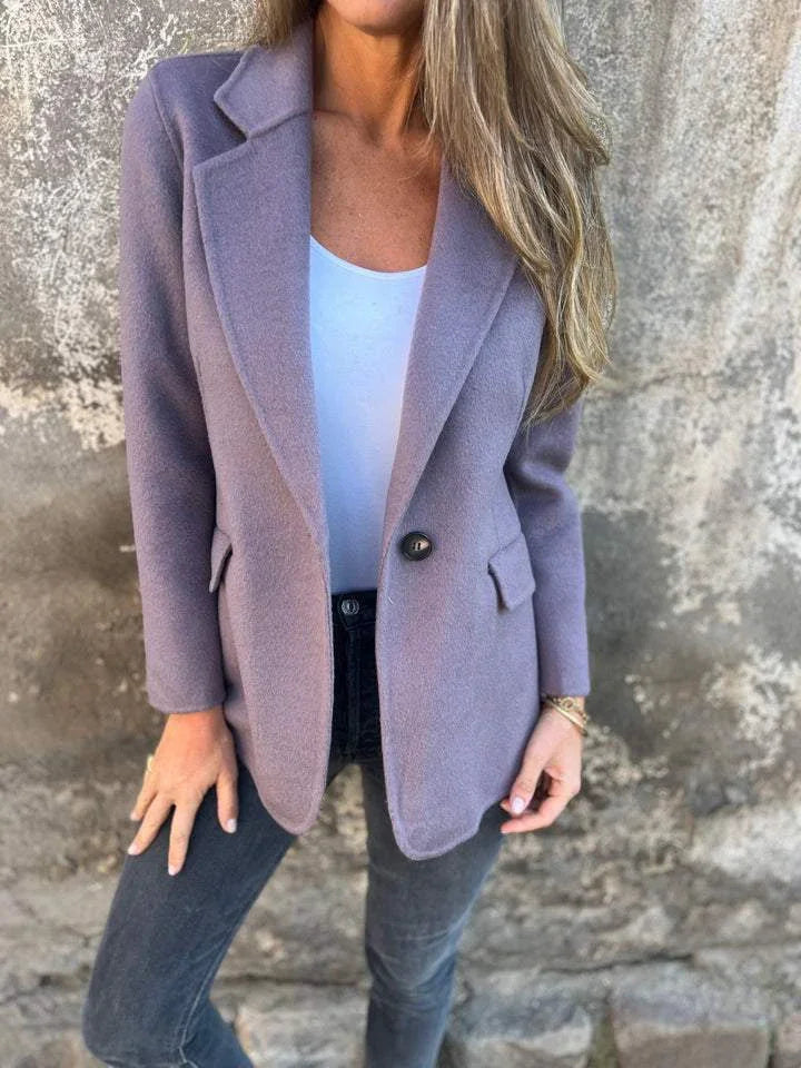 Woman wearing a lavender single-button wool blazer over a white top, paired with dark jeans, standing against a textured stone wall
