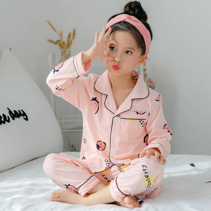 Girl in pink strawberry print pajamas and matching headband sitting on bed making hand circle gesture