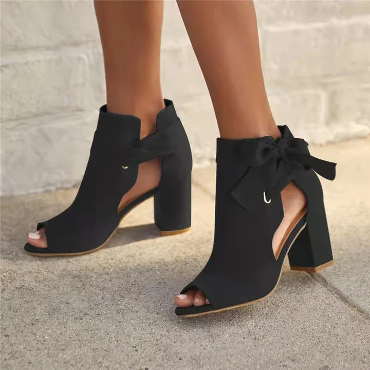 Black open-toe high heel ankle boots with cutouts and bow ties on sidewalk