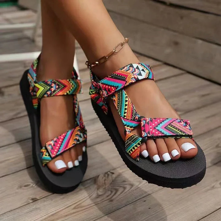Colorful patterned summer sandals with black soles worn on feet with white pedicure on wooden floor
